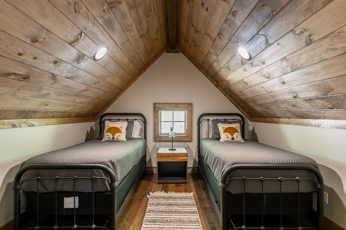 A cozy side room with two twin beds, decorative pillows, a small nightstand with a lamp, and a framed window. The warm, welcoming setting is perfect for intimate sleepovers or quiet time away from the main spaces.