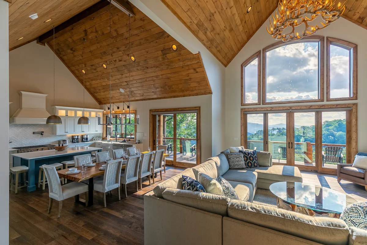 This image showcases a spacious open-concept living area featuring large windows that frame an impressive mountain view, a wooden ceiling with exposed beams, a modern kitchen island, a generous dining table, and a comfortable seating area. The natural light pours into the space, highlighting the seamless connection between the living, dining, and kitchen zones.