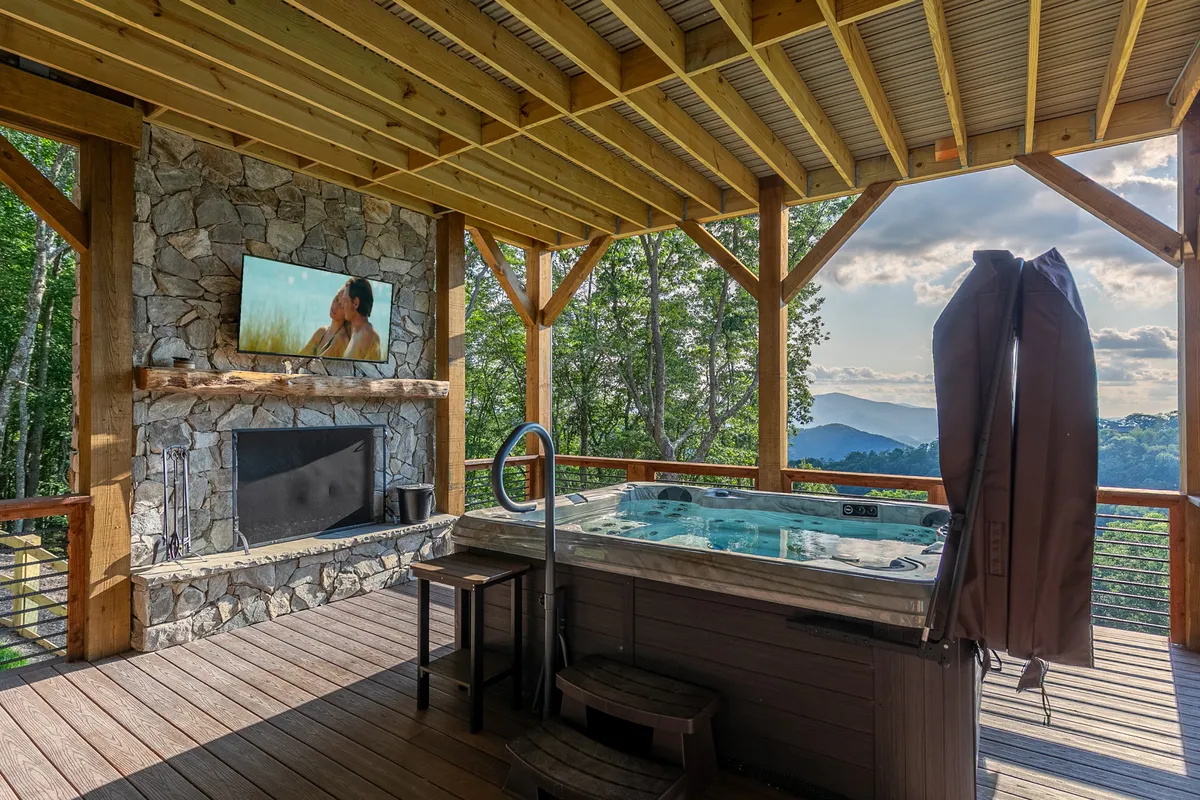 A luxurious deck scene featuring a large hot tub with mountain views, an outdoor fireplace, and a television screen. People are relaxing in the hot tub, watching movies or shows, and enjoying the tranquil natural surroundings.