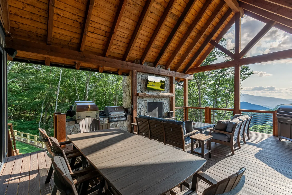 This image features a spacious wooden deck equipped with a stone fireplace, outdoor grill, large wooden dining table, comfortable seating, and expansive windows overlooking natural mountain surroundings. The scene is set for outdoor dining, barbecuing, enjoying the view, or watching TV outdoors.