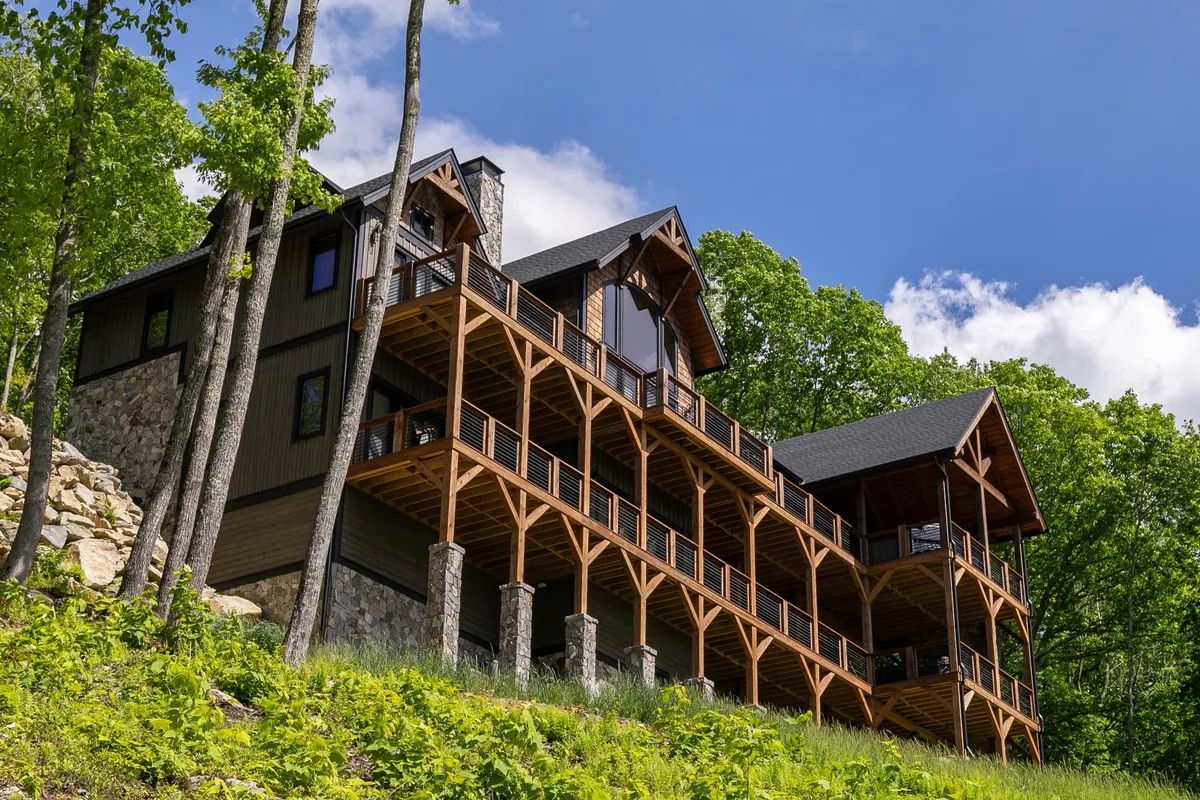 A large, inviting wooden deck wraps around the home's stone facade, nestled among tall trees. Expansive windows connect the interior to the scenic outdoors. The deck is ideal for group activities, outdoor workshops, or simply unwinding together in nature.