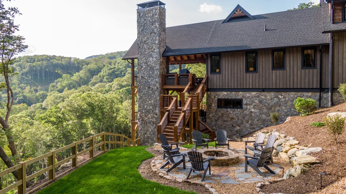 An exterior view featuring a spacious wooden deck with a stone chimney, a cozy fire pit seating area, and lush greenery surrounding the home. The setting is perfect for gathering around the fire, socializing as a family, or stargazing after a day of adventure.