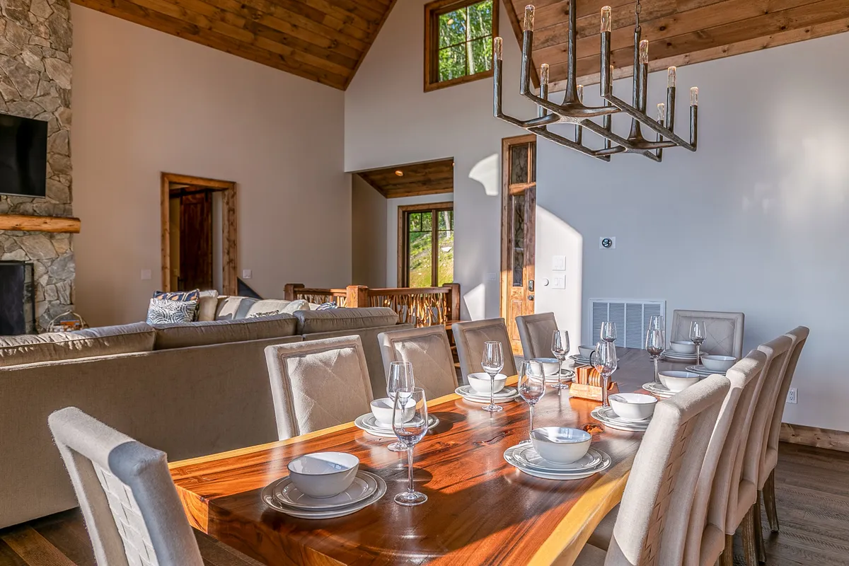 This image features an elegant dining table set for a celebration, a glowing chandelier overhead, a stone fireplace in the background, and expansive windows that let in natural light. The setting feels festive yet intimate, ideal for both special occasions and everyday gatherings.