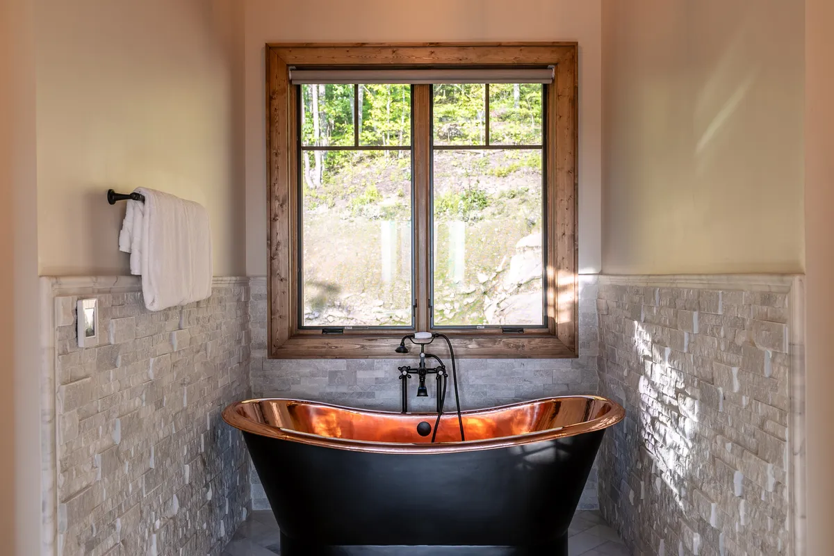 A luxurious bathroom highlighted by a gleaming copper bathtub, set against a stone wall with a large window framing views of nature. This spa-like environment invites relaxation and indulgence, echoing high-end touches.