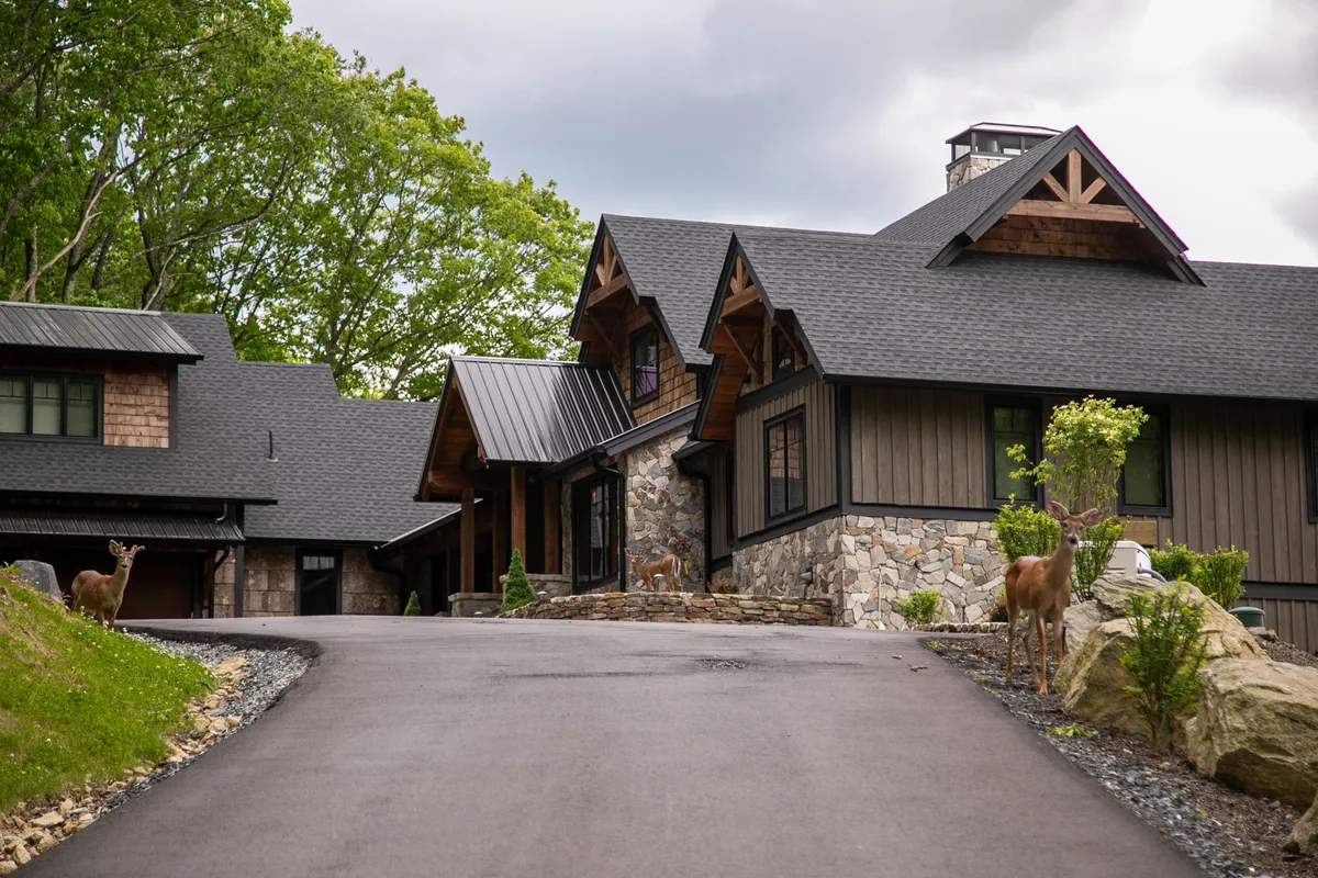 An exterior scene showing the rustic stone and wood architecture of the home, with deer grazing along a tree-lined driveway. The setting immerses guests in the natural landscape, evoking the sense of discovery and wildlife encounters just outside the door.