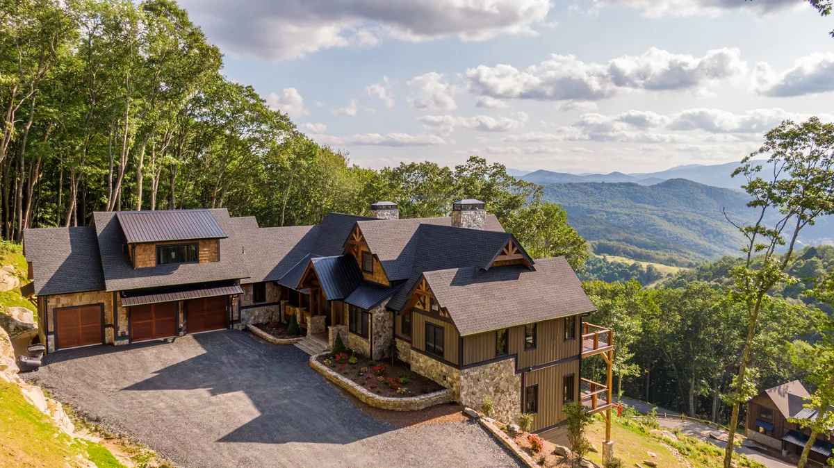The hero image showcases a luxurious, spacious cabin set amidst lush trees with sweeping mountain views in Banner Elk, North Carolina. The grand main house is the focal point, surrounded by natural beauty and an inviting, serene atmosphere. The setting highlights the property's privacy, expansive outdoor areas, and the breathtaking panorama perfect for sunsets and gatherings.