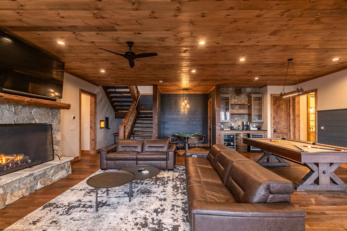 This image showcases a spacious interior featuring a stone fireplace, leather sofas, a pool table, and a wooden staircase. The room exudes a sense of comfort and fun, highlighting multiple entertainment amenities in a single view.
