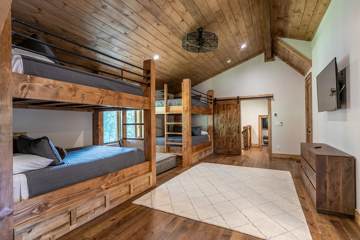 A spacious bunk room with multiple sturdy bunk beds, a wooden ceiling, a large window letting in daylight, a cozy area rug, and a wall-mounted television. The space is inviting, playful, and ideal for groups of kids or friends.