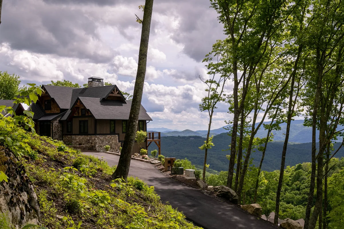 A spacious wooden deck overlooks a stunning mountain vista, bordered by lush greenery. The stone exterior of the home and large windows anchor the scene, suggesting a seamless blend of indoor comfort and outdoor beauty. The deck offers ample space for gatherings, workshops, or relaxed socializing while enjoying panoramic views.