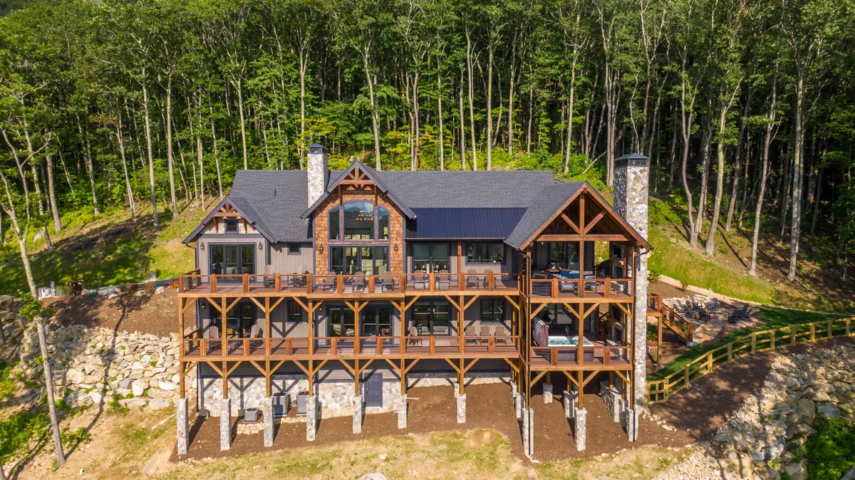A wide wooden deck built atop a sturdy stone foundation, surrounded by towering forest trees and featuring large windows that connect the indoors with the outdoors. The deck is set up for outdoor dining and family gatherings, inviting guests to relax and enjoy nature.