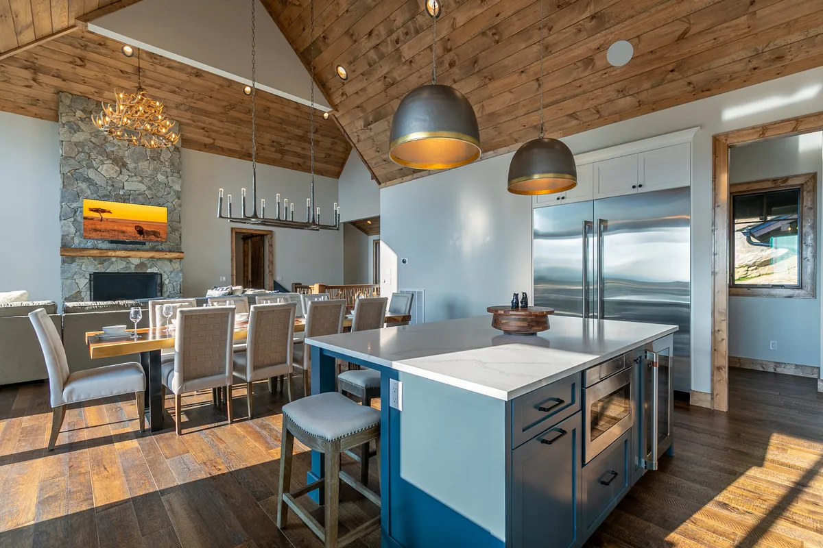 The image focuses on an expansive dining table that seats a large group, a chef-style kitchen island, a stunning stone fireplace, and a chandelier. The layout makes it easy to envision family dinners, celebrations, and casual breakfasts, all set within a warm, inviting ambiance.